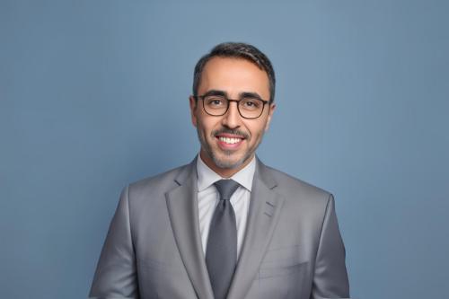 Mina Sedrak smiles at the camera. He wears a grey suit and dark grey tie.