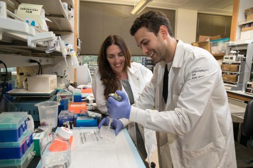 April Pyle smiles and looks on while a trainee in her lab uses a pipet. 