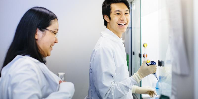 A UCLA researcher stands smiling with a pipet while another looks on.